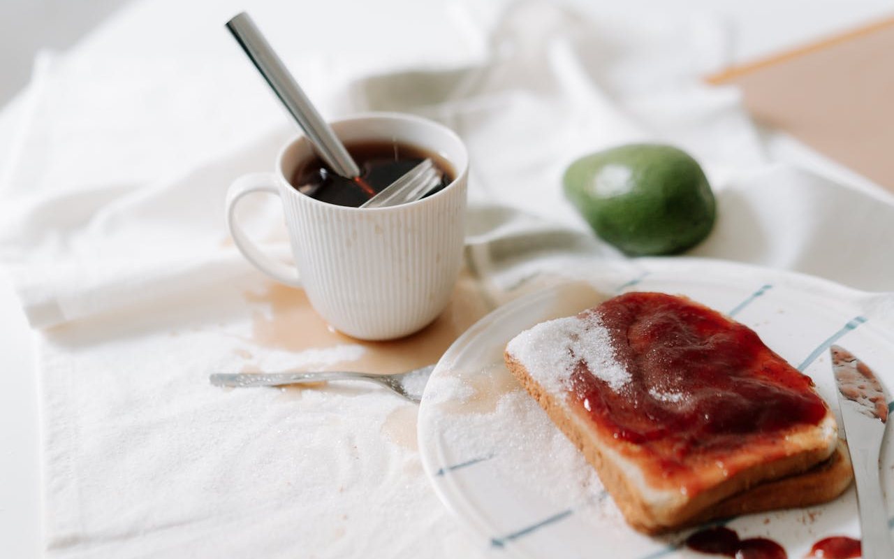 Bela tkanina na kojoj se nalazi šolja kafe ispod koje je mrlja od iste, a pored koje je avokado i tanjir sa hlebom namazanim marmeladom.