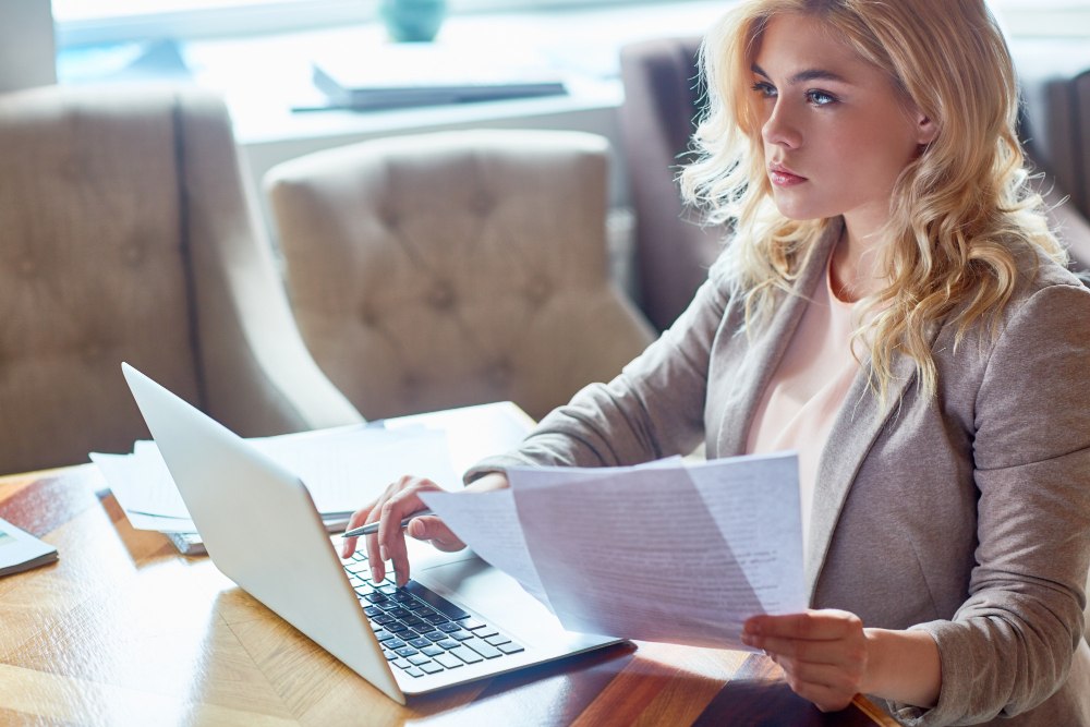 zena sa papirima u ruci ispred otvorenog laptopa